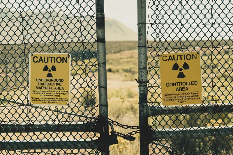 Maschendrahtzaun mit zwei gelben Warnschildern „Caution“ vor einer kargen Landschaft, das Tor mit einer Kette verschlossen
