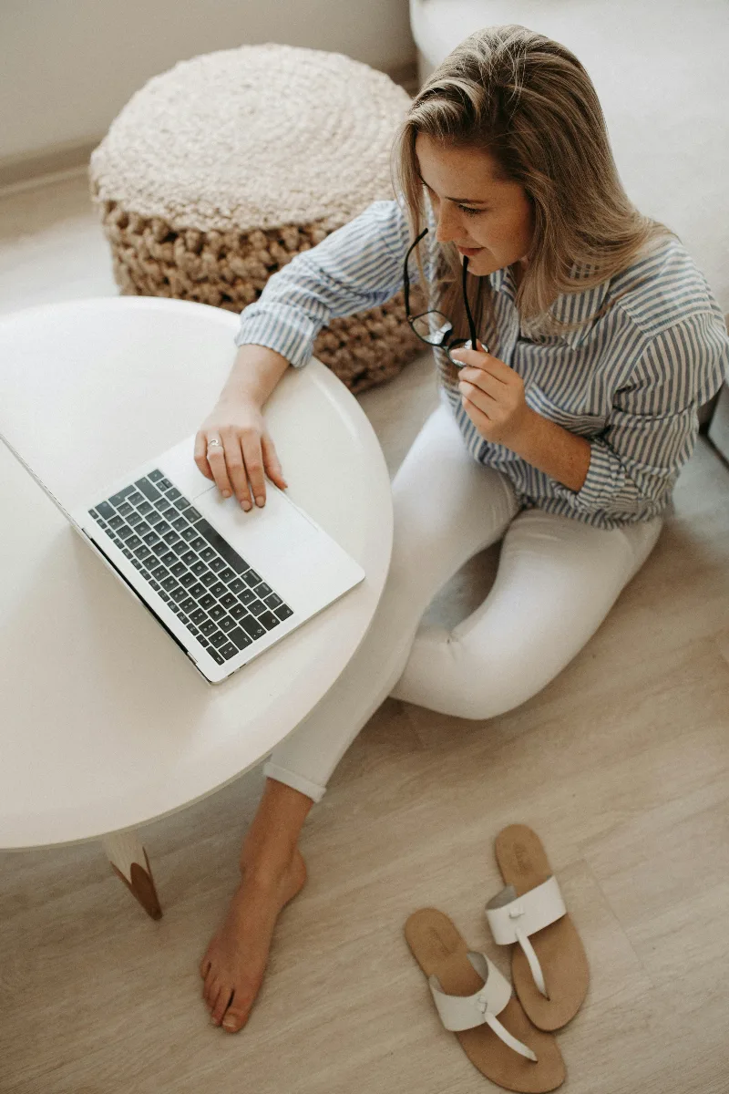 Frau sitzt mit dem Laptop an einem niedrigen Tisch und arbeitet, Brille in der Hand.