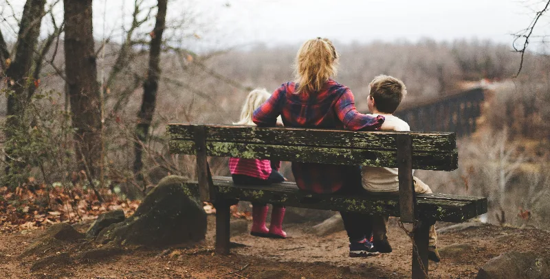 Eine Mutter sitzt mit zwei Kindern auf einer Bank am Waldrand, von hinten fotografiert.